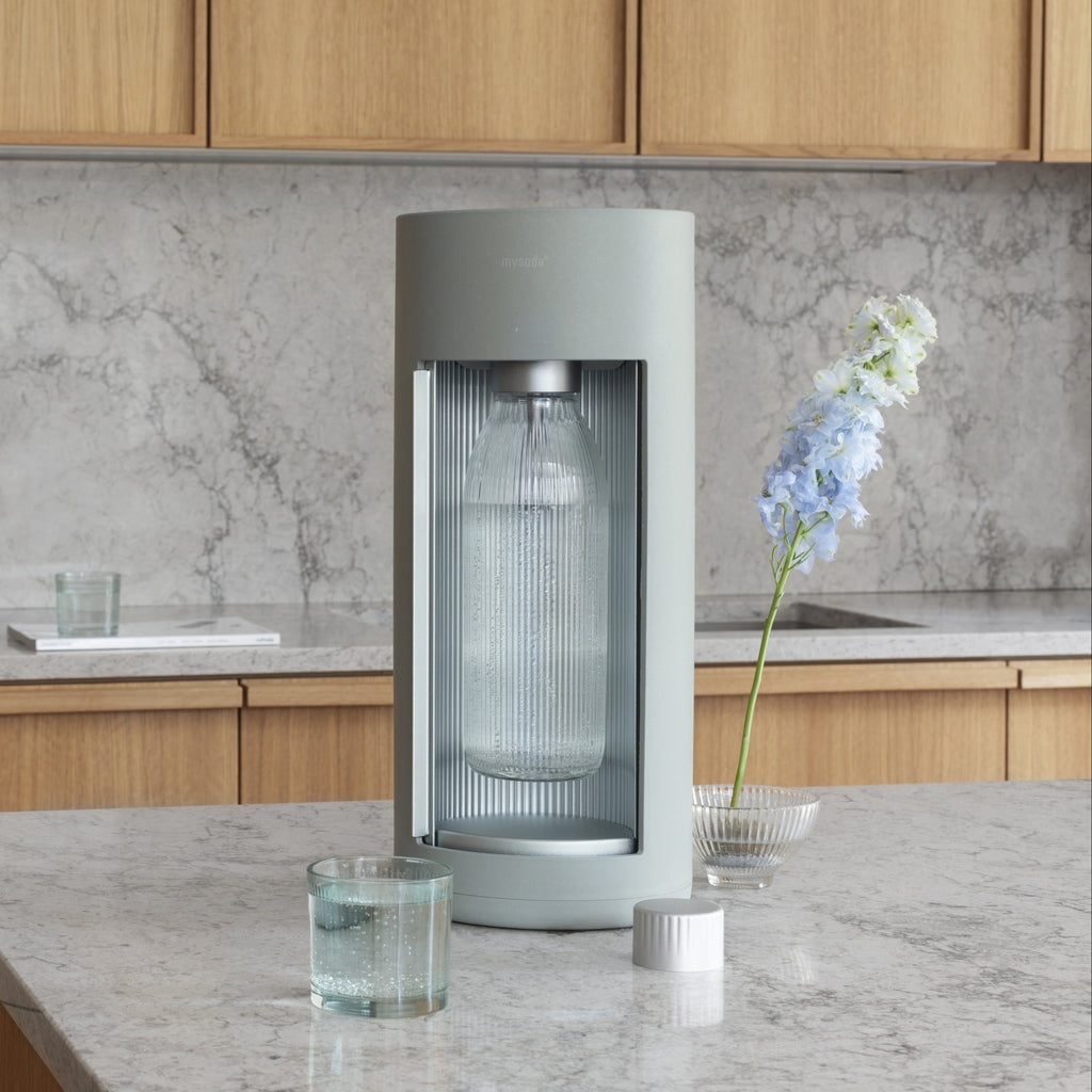 A pigeon coloured Glassy soda maker in a kitchen with gray marble counters and cupboards in light wood