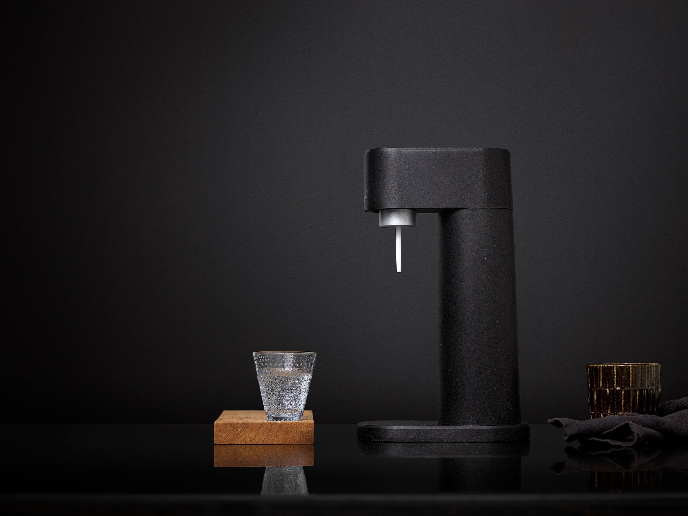 A black Woody soda maker and glass of sparkling water in an all-black studio setting
